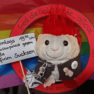 Plüschmaus mit rotem Irokesenschnitt, schwarzer Jacke mit Ketten und Aufnäher '161', hält Schild mit Text 'montags 19:00 Uhr Kulturpalast gegen die freien Sucksen', vor Regenbogenflagge und rotem Kreis mit Text 'fred.dielinkesocke_official'