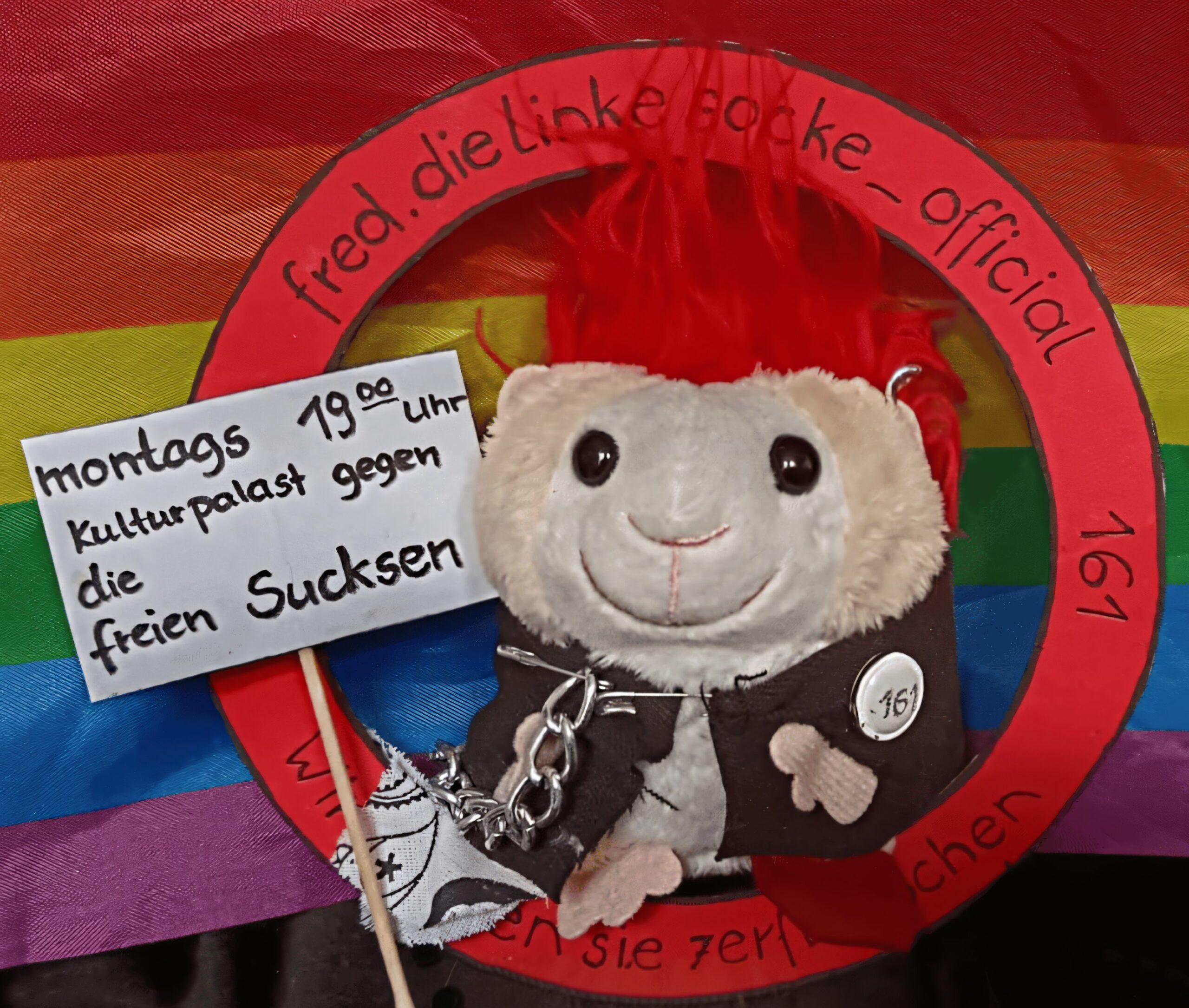 Plüschmaus mit rotem Irokesenschnitt, schwarzer Jacke mit Ketten und Aufnäher '161', hält Schild mit Text 'montags 19:00 Uhr Kulturpalast gegen die freien Sucksen', vor Regenbogenflagge und rotem Kreis mit Text 'fred.dielinkesocke_official'