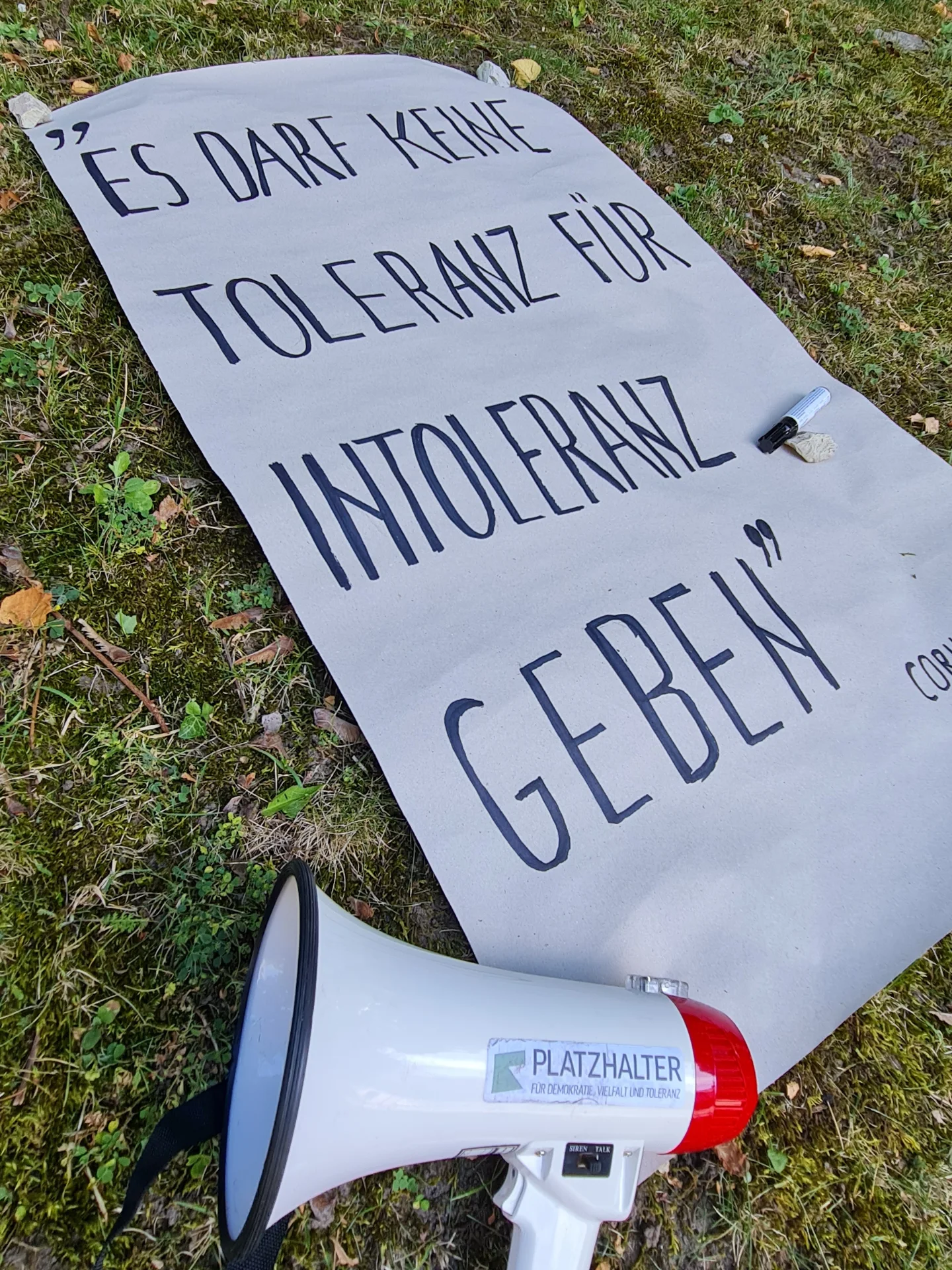 Weißes Plakat auf grüner Wiese mit schwarzer Schrift: 'Es darf keine Toleranz für Intoleranz geben' - Zitat. Auf dem Plakat liegt ein schwarzer Permanentmarker. Neben dem Plakat steht ein weißes Megafon mit rotem Griff und dem Aufkleber 'PLATZHALTER für Demokratie'.